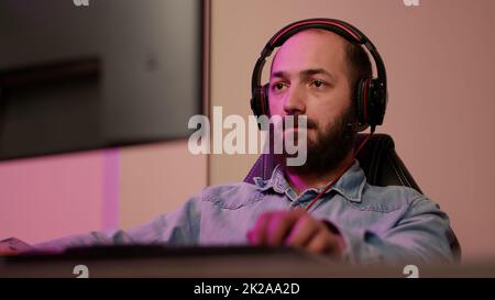 Handheld shot of man with gaming headset looking at computer screen and talking to team members while streaming gameplay. Gamer playing multiplayer online action game using professional pc setup. Stock Photo
