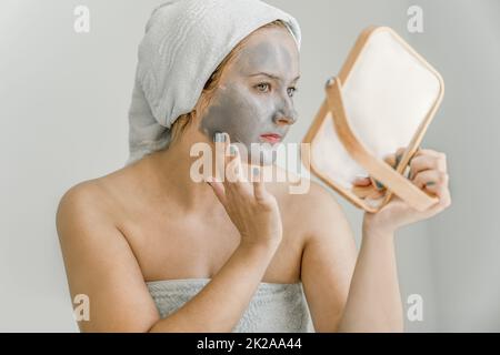 Young woman puts gray cosmetic clay mask on face while looking in mirror, her hair and body wrapped in towel, Side view. Close-up Stock Photo