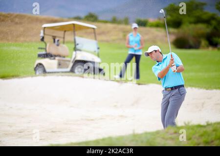 In and quickly out of a bunker. A young male golfer chipping his ball out of a bunker while his female partner looks on from the fairway. Stock Photo