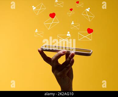 female hand holds a smartphone with flying out letters on a yellow background. Social media addiction, receiving correspondence and appraisal. Stock Photo