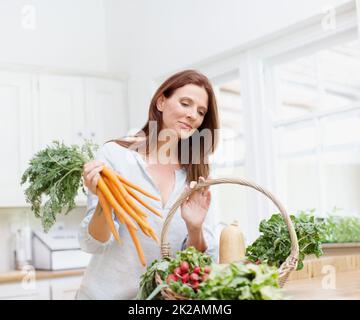 All the right ingredients for a nice minestrone soup. A gorgeous woman holds a bunch of carrots while looking into her basket of vegetables in her kitchen. Stock Photo