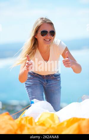 Jump success. A beautiful young woman giving two thumbs up outdoors with a parachute in the foreground. Stock Photo