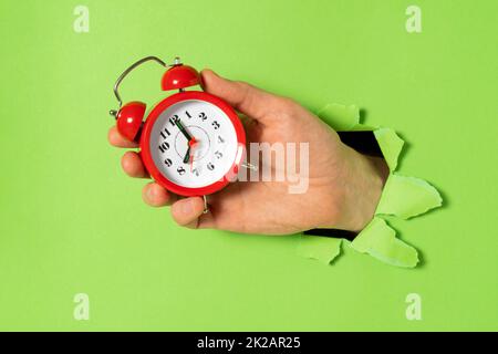 Hand with alarm clock sticks out of a torn hole in paper Stock Photo