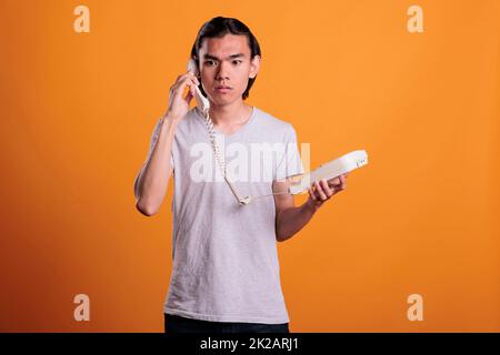 Young scared man talking on landline phone. Confused asian teen chatting, holding retro telephone, frightened person with terrified facial expression answering call on orange background Stock Photo