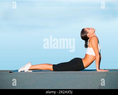 Easing her back muscles into a stretch. A young woman doing yoga outdoors against a blue sky. Stock Photo