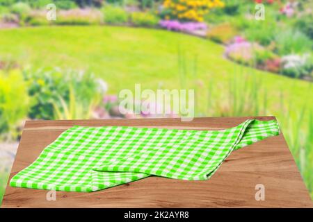 Empty table product. Empty wooden deck table covered with a green white checkered tablecloth over abstract blurred natural backdrop. Space for your food and product display montage. Stock Photo