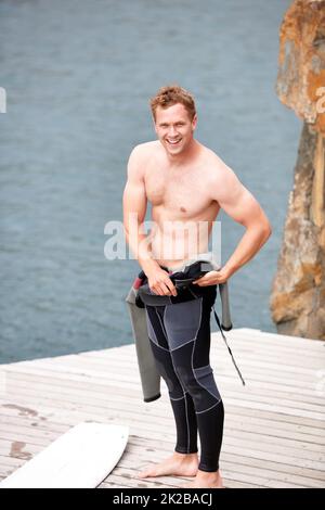 Preparing for an awesome day. Young man putting on his wetsuit before engaging in some watersports. Stock Photo