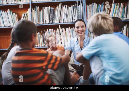 Learning together. A pretty teacher sitting with her excited students in the library. Stock Photo