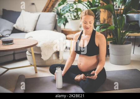 Young happy and cheerful beautiful pregnant woman chating to family and friends on mobile phone while staying fit, sporty and active on her maternity leave. Pregnancy, yoga concept Stock Photo