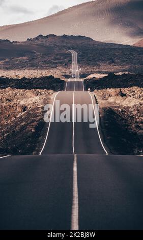 Endless road on a volcano in Timanfaya National Park in Lanzarote in the Canary Islands with a continuous line, black volcanic rocks on the side and volcanoes in mist in background. Stock Photo