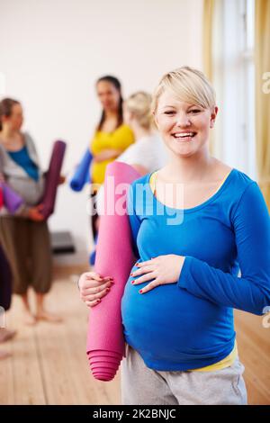 Keeping healthy for my little one. A young blonde pregnant woman in a gym holding an exercise mat with a group of women in the background. Stock Photo