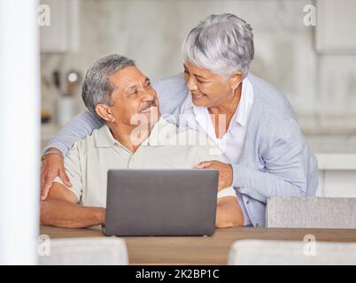 Happy elderly couple streaming on laptop, hug and bonding while relax in kitchen at home. Senior man and woman indoors, enjoy retirement and Stock Photo