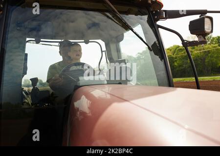 Work begins early on the farm. Shot of a young boy sitting with his dad inside the cab of a modern tractor. Stock Photo