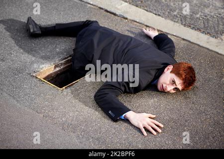 There are many obstacles on the road to success. A young businessman lying on the ground with his leg stuck in a drain. Stock Photo