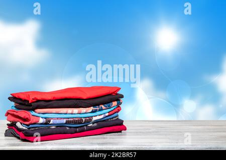 Stack colorful clothes. Closeup of a pile of colorful t-shirts or shirts on a bright table against abstract blurred sunny blue sky background. Space for your product display montage. Summer fashion. Stock Photo