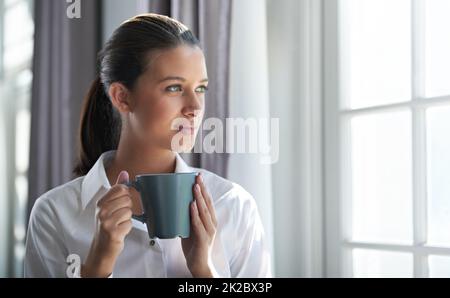 Beginning of the business day. Shot of a young businesswoman enjoying a cup of coffee while standing next to a windown. Stock Photo