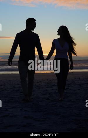 The best thing about sunsets is watching them with you. Silhouette shot of a young couple walking on the beach. Stock Photo