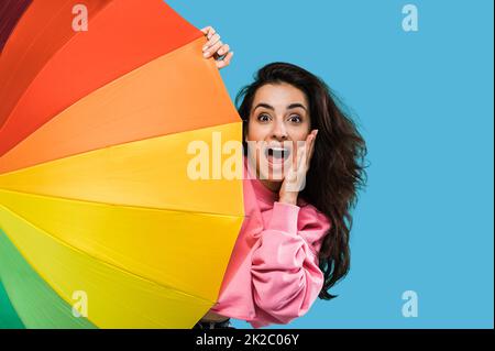 Shocked excited caucasian beautiful long haired young woman in pink sweater, peeking out from behind colorful umbrella, looking at camera in amazement, standing over isolated blue background Stock Photo