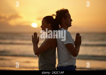 Soul experience at sunset. Shot of a couple doing yoga on the beach at sunset. Stock Photo