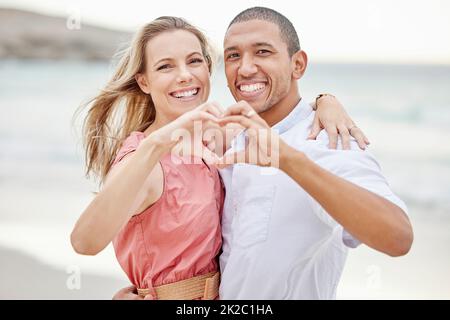 Happy interracial couple show heart sign with their hands and enjoying fresh air on vacation at the beach while bonding. Portrait of couple hugging at Stock Photo