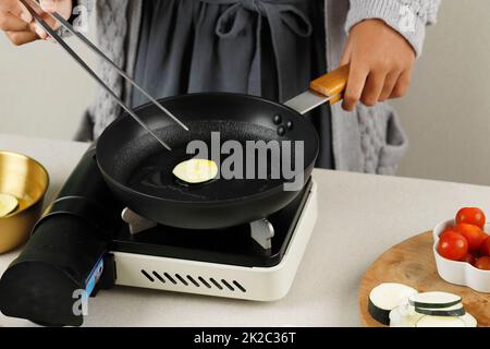 Cooking Hobak Jeon, Korean Zucchini Pancake in the Kitchen, Put Sliced Zucchini with Egg Batter on Black Pan. Female Hand Hold Stainless Tongs Stock Photo