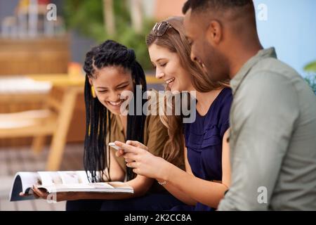 Print versus digital in the student world. Three friends reading from a book and a cellphone while sitting outdoors. Stock Photo