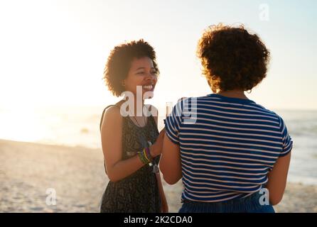 You really make me laugh. Cropped shot of two attractive young women standing on the beach and enjoying a drink together during the day. Stock Photo
