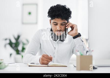 Can you repeat that please. Shot of a young businessman writing notes while talking on a cellphone in an office. Stock Photo