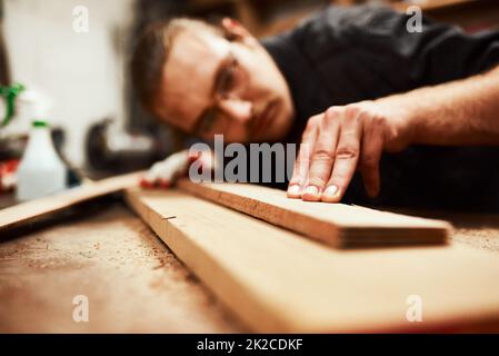 Thats the straightest its going to get. Cropped shot of a focused young male carpenter blowing dust off of a piece of wood after sanding it inside of his workshop during the night. Stock Photo