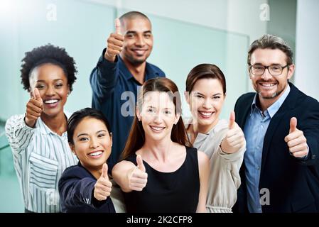 Stay positive, work hard and make it happen. High angle portrait of a group of businesspeople standing together and showing a thumbs up in the office. Stock Photo