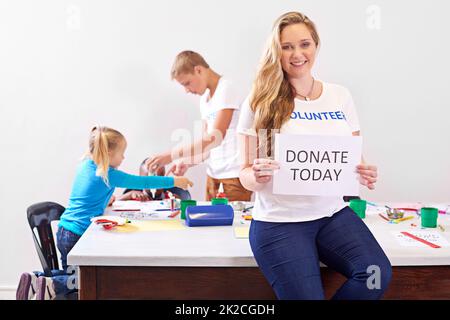 Your help is always needed. Portrait of a volunteer holding up a donate today sign while working with little children. Stock Photo