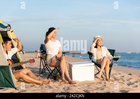 Happy friends with drinks in camp and laughing together in holidays on sand beach near camping tent vacation time at sunset Stock Photo
