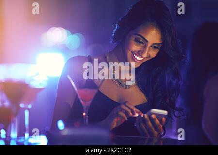 Get here now, this party is great. Shot of a woman using her cellphone while sitting at a table in a nightclub. Stock Photo