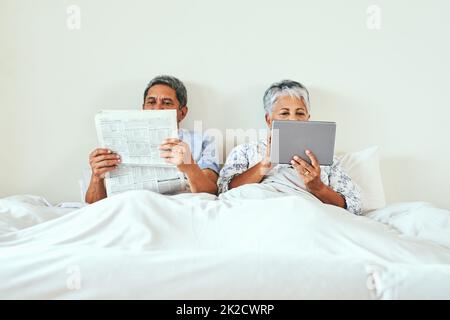 Keeping up with the world the best way they can. Shot of a relaxed mature couple lying in bed while browsing on a digital tablet and reading the newspaper at home in the morning. Stock Photo