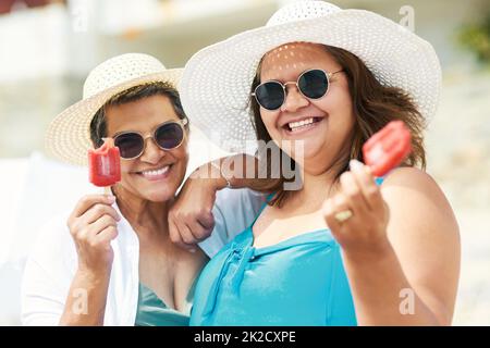 The perfect summer cool down. Shot of two mature friends standing together and enjoying ice cream while bonding on the beach during the day. Stock Photo