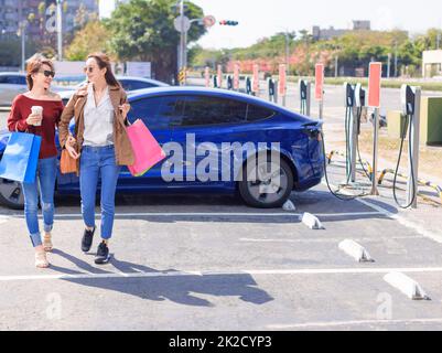 Happy  young woman standing on city parking near electric car, charging automobile battery from small city station, holding shopping bags and walking together Stock Photo