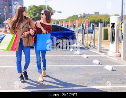 Happy  young woman standing on city parking near electric car, charging automobile battery from small city station, holding shopping bags and walking together Stock Photo