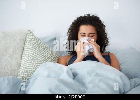 I definitely cant go to work with this cold. Shot of an attractive young woman feeling sick and blowing her nose while in bed in the morning. Stock Photo