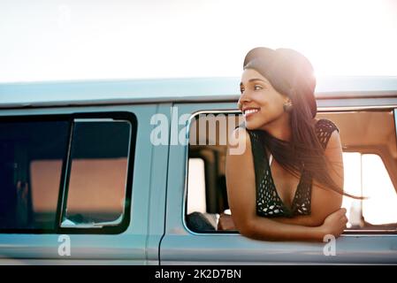 Off to see whats out there. Shot of a happy young woman leaning out of the window during a road trip. Stock Photo