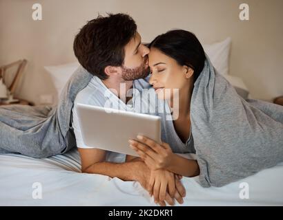 Couple, love and tablet for entertainment while lying in bed with a kiss on forehead while watching a movie, internet browsing and streaming service Stock Photo