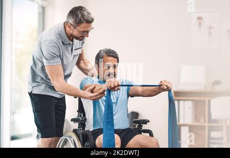 This exercise is also great for maintaining good balance. Shot of a senior man in a wheelchair exercising with a resistance band along side his physiotherapist. Stock Photo