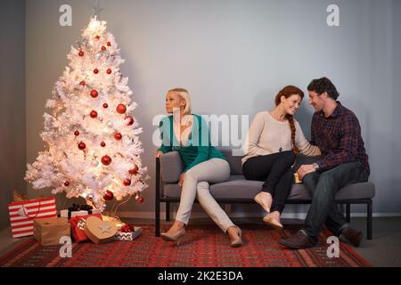 Someones getting their tinsel in a tangle. Shot of a couple bonding while their friend sits alone by the Christmas tree. Stock Photo