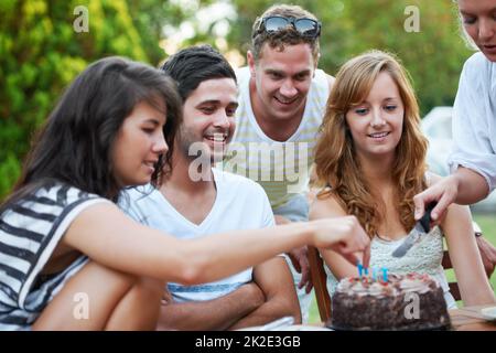 Birthdays are all about sharing. Smiling young teen friends celebrating someones birthday and cutting some birthday cake. Stock Photo