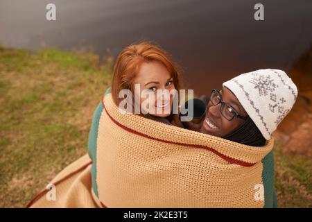 As close as two best friends can get. A high angle portrait of two happy young women huddled under a blanket next to a lake in winter. Stock Photo