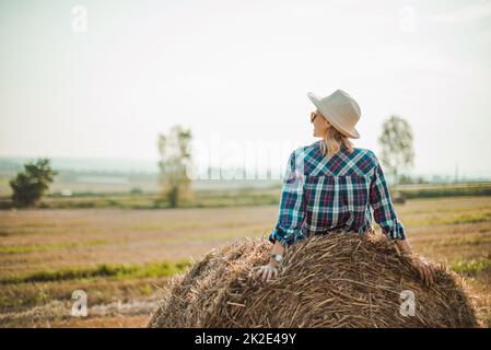 Woman in hat sitting on round bales in the field, enjoying good weather. Stock Photo