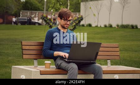 Successful busy caucasian young man afro guy student male freelancer programmer designer in wireless headphones sitting on terrace in city in park typing laptop chat working remotely e-learning online Stock Photo