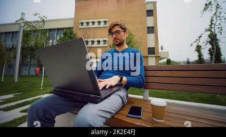 Working from park. Handsome student studying on computer at campus outdoors and drinks coffee. Stock Photo