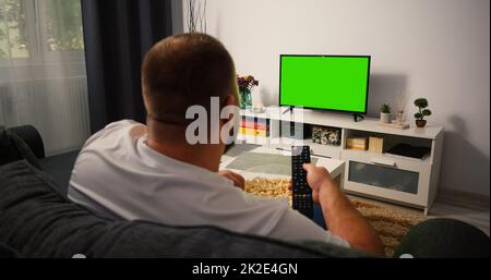 Back view Man sitting on the couch in the living room Watching TV with Green Screen and Changing Channels with Remote Control in his hand. Chroma Key TV in focus. Stock Photo