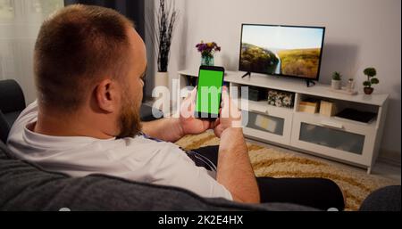 Smartphone Vertical Green Screen for Copy Space Chroma Key Mockup. Back over the shoulder view man watching Video movie on couch Home. Shot Over the Shoulder Green Screen phone. Stock Photo