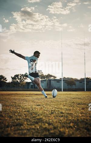 His technique is unmatched. Full length shot of a handsome young rugby player kicking a ball on the field during the day. Stock Photo